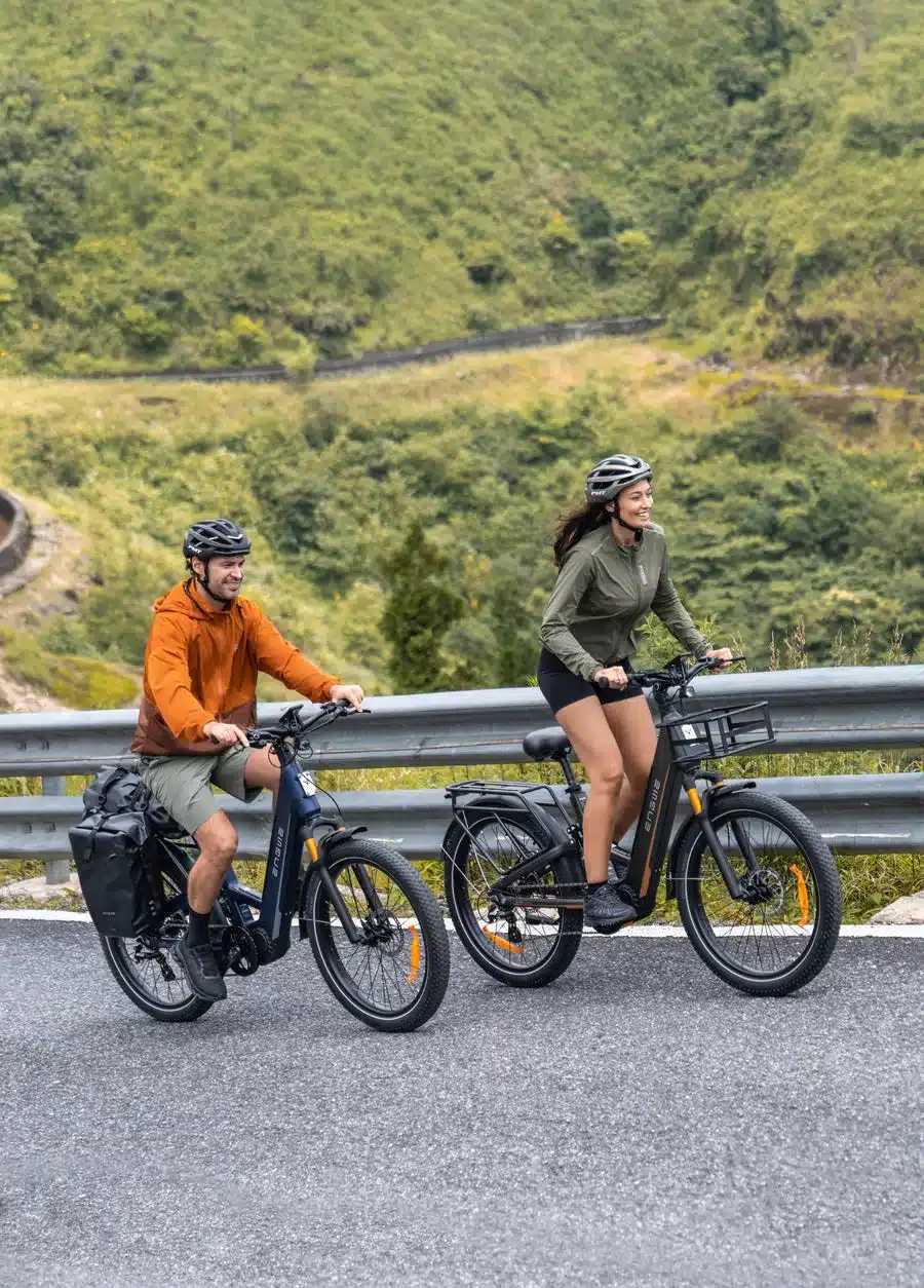 Mann und Frau fahren auf ENGWE E26 3.0 Pro E-Bikes auf einer asphaltierten Straße mit grünen Hügeln im Hintergrund.