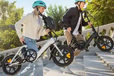 Ein Mann und eine Frau tragen jeweils ein weißes und ein schwarzes Fiido D3 Pro Mini E-Bike auf einer Steintreppe in der Sonne.
