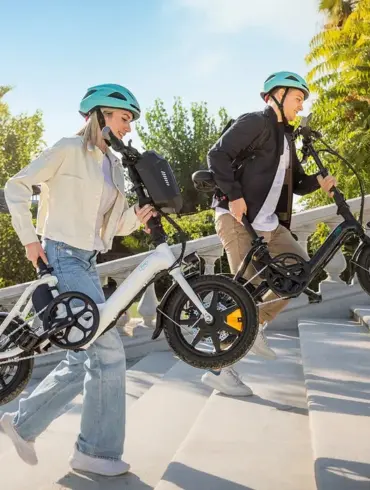 Ein Mann und eine Frau tragen jeweils ein weißes und ein schwarzes Fiido D3 Pro Mini E-Bike auf einer Steintreppe in der Sonne.