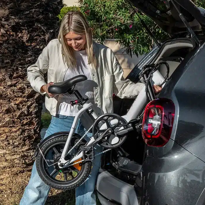 Frau mit hellem Hemd und Jeans verstaut ein weißes Fiido D3 Pro Mini-Klapp-E-Bike in den offenen Kofferraum eines dunklen Autos neben einem Baum.