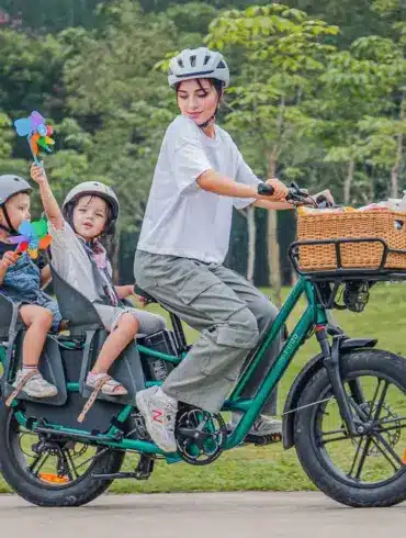 Eine Frau fährt ein grünes Fiido T2 Lastenrad mit zwei Kindern auf Kindersitzen im Park.