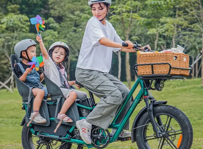 Eine Frau fährt ein grünes Fiido T2 Lastenrad mit zwei Kindern auf Kindersitzen im Park.