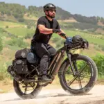 Mann in schwarzer Kleidung und Helm fährt ein schwer beladenes schwarzes Fiido Titan E-Bike mit breiten Fat-Reifen auf einem Feldweg.