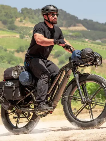 Mann in schwarzer Kleidung und Helm fährt ein schwer beladenes schwarzes Fiido Titan E-Bike mit breiten Fat-Reifen auf einem Feldweg.