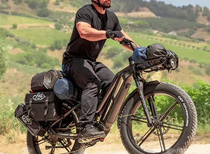 Mann in schwarzer Kleidung und Helm fährt ein schwer beladenes schwarzes Fiido Titan E-Bike mit breiten Fat-Reifen auf einem Feldweg.