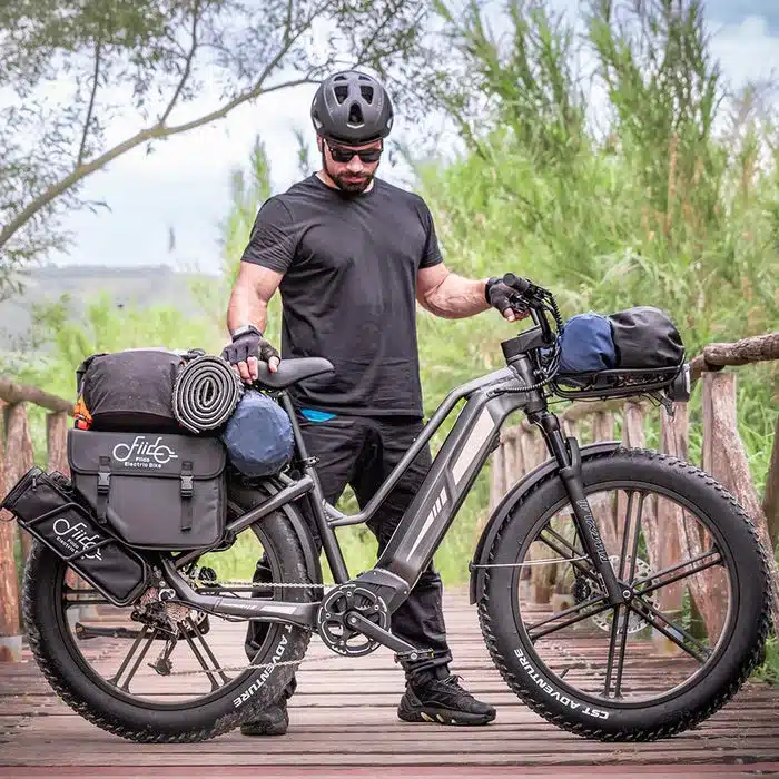 Ein Mann in schwarzer Kleidung und Helm steht neben einem grauen Fiido Titan E-Bike, das mit Gepäcktaschen, einer Rolle und einer Isomatte beladen ist. Das Fahrrad hat breite Fat-Reifen und steht auf einem Holzsteg inmitten von Grün.