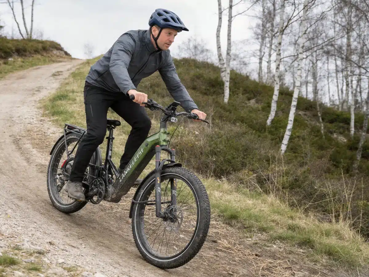 Ein Mann in dunkler Kleidung und Helm fährt ein grünes E-Bike mit breiten Reifen auf einem schmalen Feldweg bergab. Hinter ihm ist ein Wald mit kahlen Bäumen und Buschwerk.