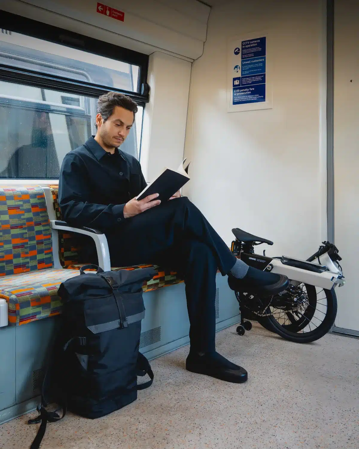 Ein Mann sitzt auf einem Sitz in einem Zug und liest ein Buch. Neben ihm auf dem Boden steht ein zusammengeklapptes weißes und schwarzes Lemmo Zero Faltrad, daneben ein schwarzer Rucksack.