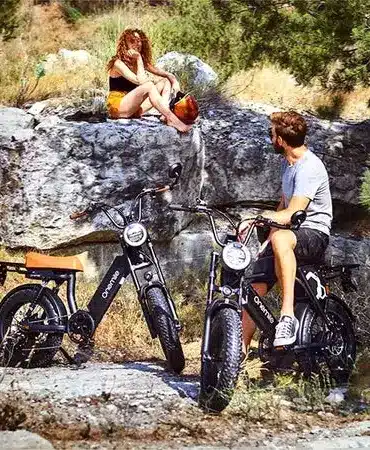 Zwei Onemile Scrambler S Pro E-Bikes stehen in einer felsigen, bewachsenen Landschaft. Eine Frau sitzt auf einem Felsen und lehnt sich zurück, während ein Mann auf einem der E-Bikes sitzt und in die Ferne blickt.