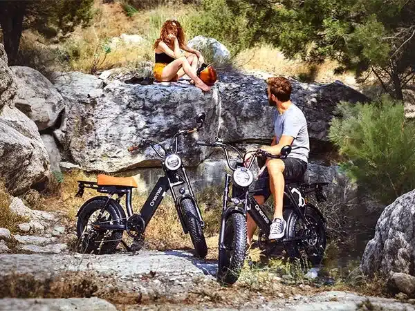 Zwei Onemile Scrambler S Pro E-Bikes stehen in einer felsigen, bewachsenen Landschaft. Eine Frau sitzt auf einem Felsen und lehnt sich zurück, während ein Mann auf einem der E-Bikes sitzt und in die Ferne blickt.