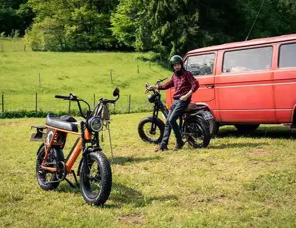 Orangefarbenes Onemile Scrambler V Pro E-Bike steht im Vordergrund, ein weiteres schwarzes E-Bike neben einem roten VW Bulli, im Hintergrund eine grüne Wiese und Bäume.