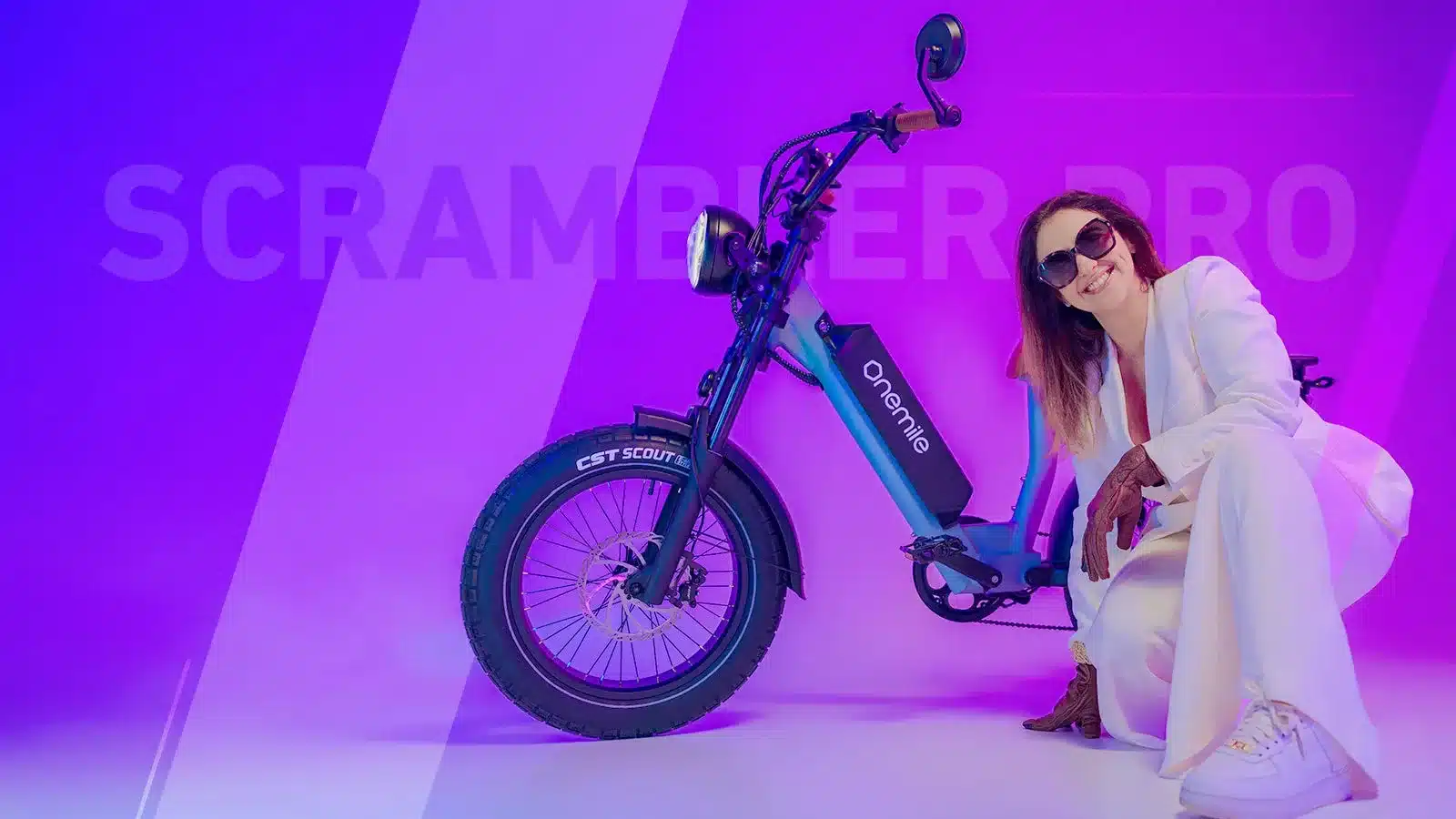 Ein blaues Onemile Scrambler V Pro E-Bike steht neben einer Frau in einem weißen Hosenanzug und Sonnenbrille, mit dem Schriftzug
