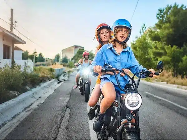Zwei Frauen auf einem Onemile Scrambler V Pro E-Fatbike fahren auf einer asphaltierten Straße, eine hinter der anderen. Sie tragen bunte Helme und sehen nach vorne.