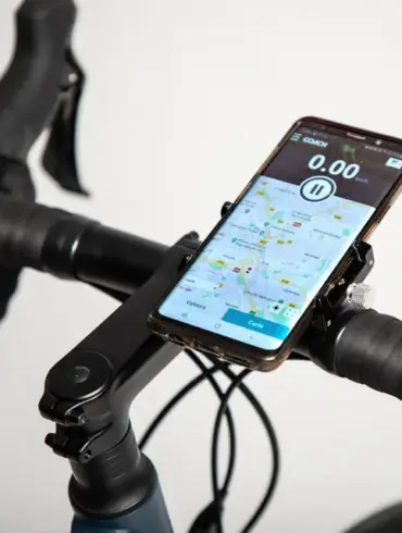 Detailansicht einer schwarzen Metall-Smartphonehalterung, befestigt an einem Fahrradlenker. Ein Smartphone mit Karten-App und Tachometer ist in der Halterung angebracht.