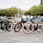 Drei stilvolle E-Bikes im Retro-Chopper-Look, das Modell Ruffian, stehen nebeneinander auf einer Holzpromenade mit Wasser und Bäumen im Hintergrund.
