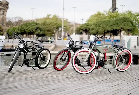 Drei stilvolle E-Bikes im Retro-Chopper-Look, das Modell Ruffian, stehen nebeneinander auf einer Holzpromenade mit Wasser und Bäumen im Hintergrund.