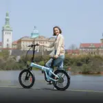 Frau auf einem hellblauen ADO Air 20 Pro Elektro-Faltrad neben einem See, mit historischer Stadtarchitektur im Hintergrund.