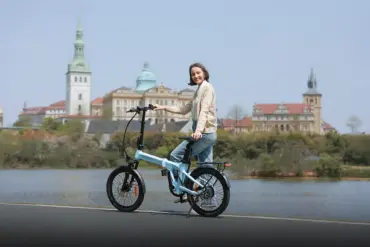 Frau auf einem hellblauen ADO Air 20 Pro Elektro-Faltrad neben einem See, mit historischer Stadtarchitektur im Hintergrund.
