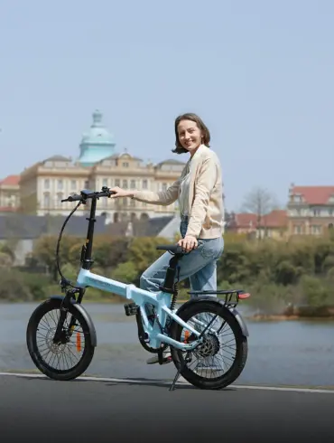 Frau auf einem hellblauen ADO Air 20 Pro Elektro-Faltrad neben einem See, mit historischer Stadtarchitektur im Hintergrund.