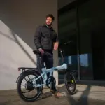 Ein Mann in dunkler Jacke steht neben einem hellblauen ADO Air 20 Ultra 2026 E-Faltrad im Freien.