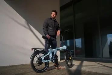 Ein Mann in dunkler Jacke steht neben einem hellblauen ADO Air 20 Ultra 2026 E-Faltrad im Freien.