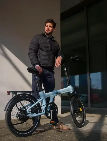 Ein Mann in dunkler Jacke steht neben einem hellblauen ADO Air 20 Ultra 2026 E-Faltrad im Freien.