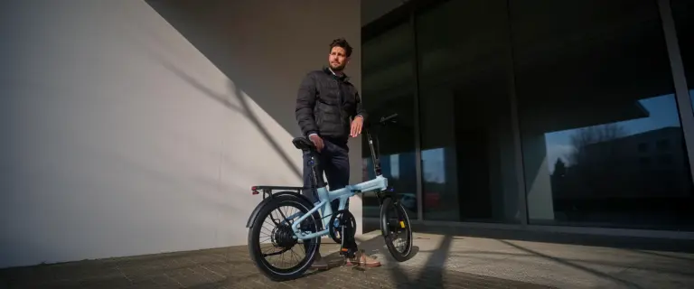 Ein Mann in dunkler Jacke steht neben einem hellblauen ADO Air 20 Ultra 2026 E-Faltrad im Freien.