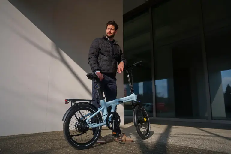 Ein Mann in dunkler Jacke steht neben einem hellblauen ADO Air 20 Ultra 2026 E-Faltrad im Freien.