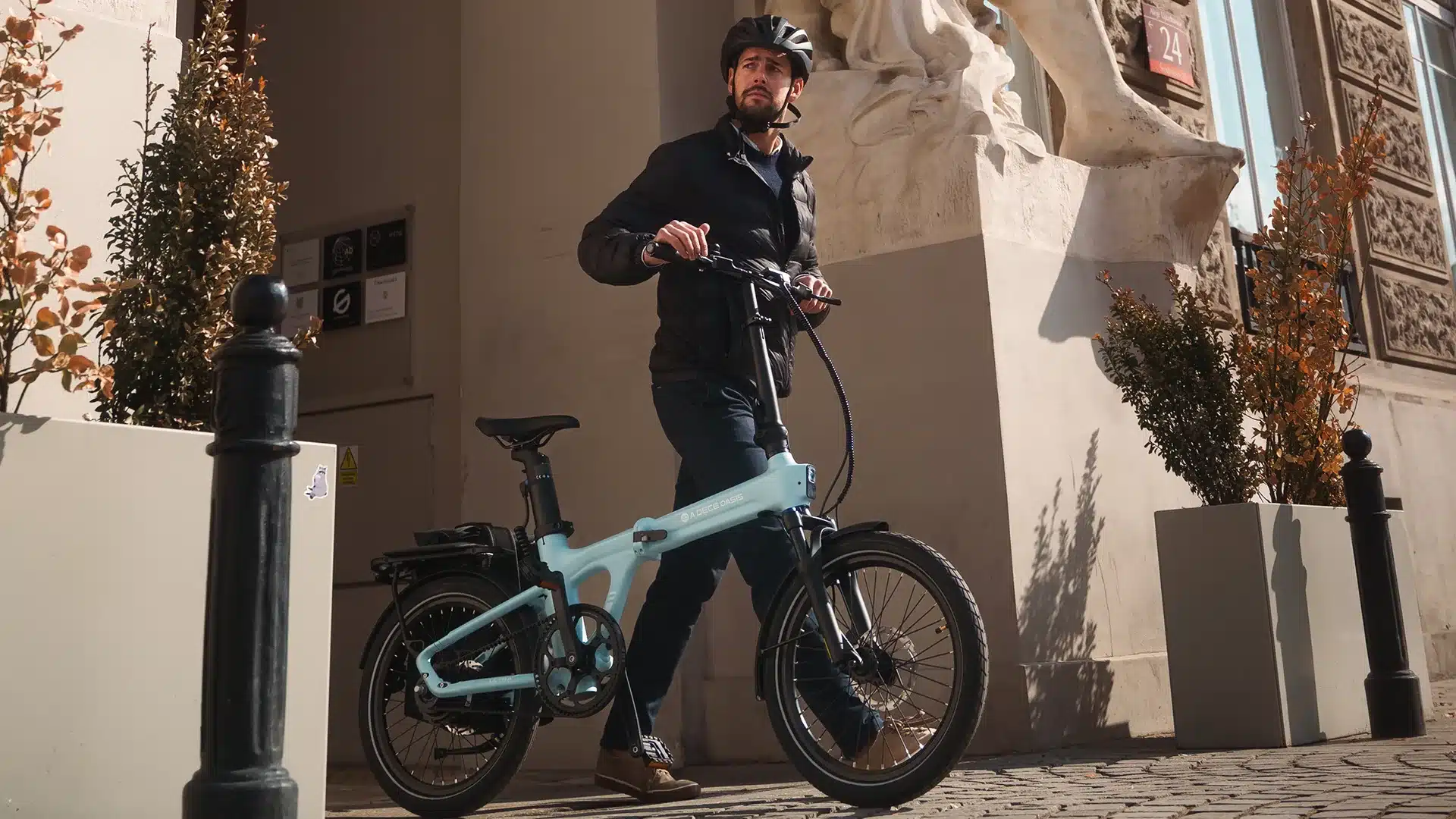 Mann mit einem hellblauen ADO Air 20 Ultra 2026 Elektro-Faltrad, das er am Lenker festhält, während er auf einem gepflasterten Bürgersteig steht.