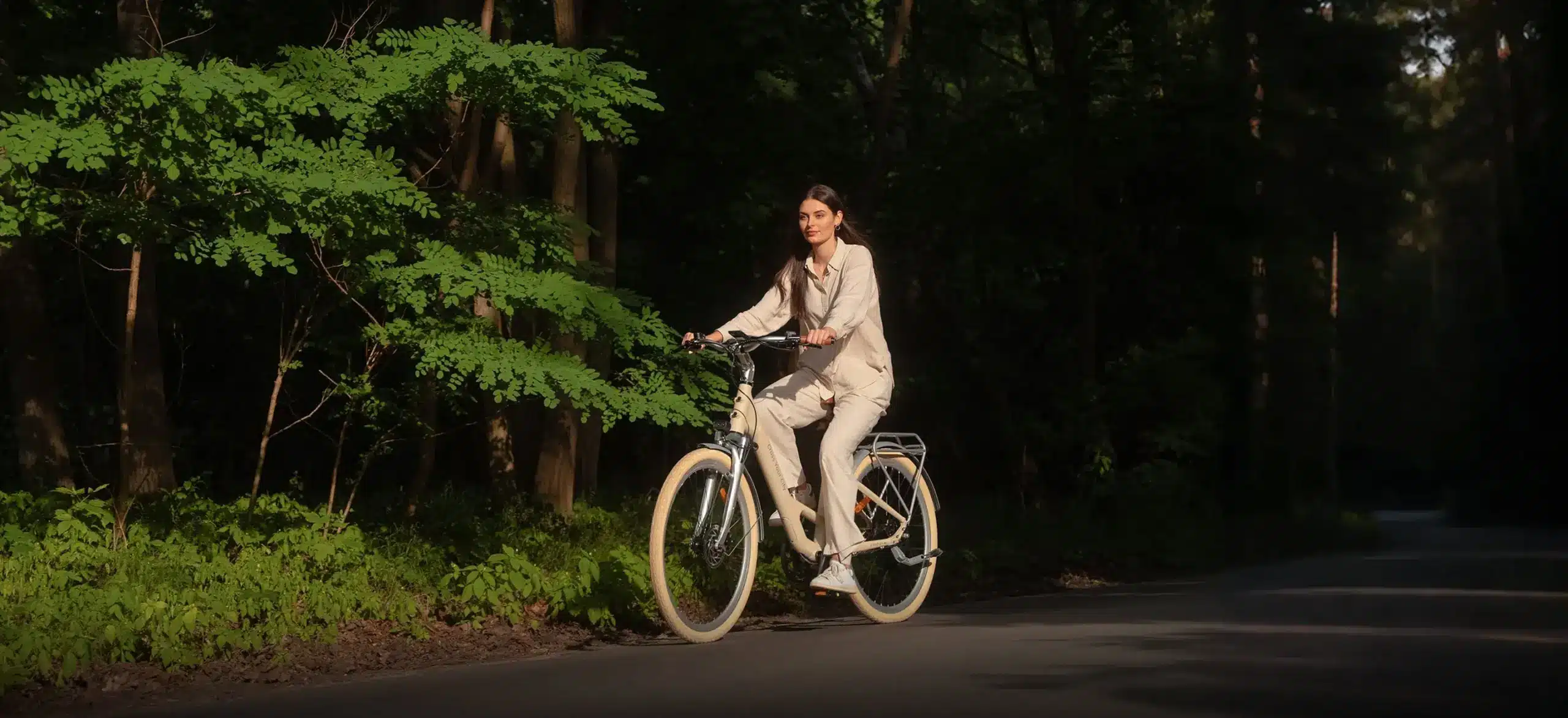 Eine Frau fährt ein beiges ADO Air 28 Pro E-Bike auf einer Straße in einem bewaldeten Gebiet.