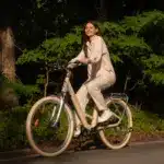 Eine lächelnde Frau sitzt auf einem beigen ADO Air 28 Pro E-Bike und fährt auf einem Weg im Freien.