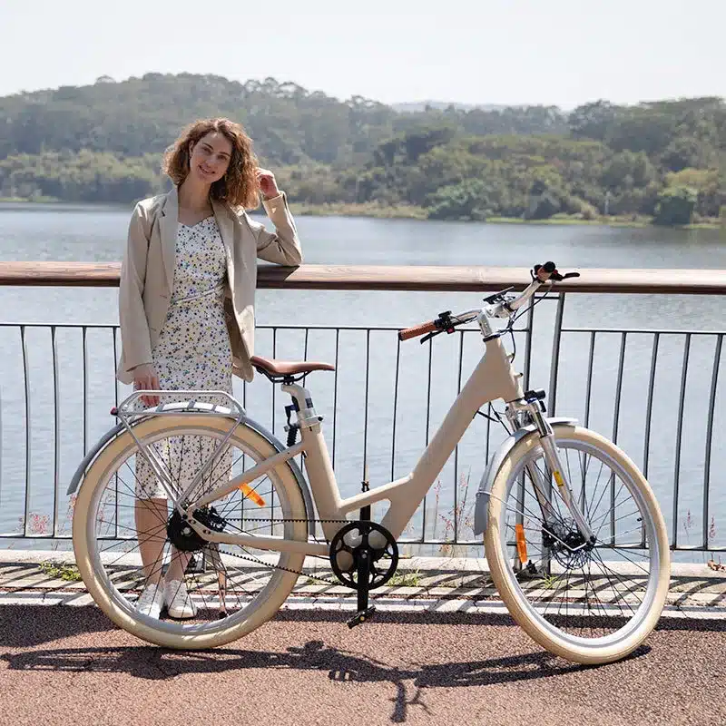 Eine Frau lehnt lässig an einem sandfarbenen ADO Air 28 Pro E-Bike, das an einem Geländer am Ufer eines Sees steht.