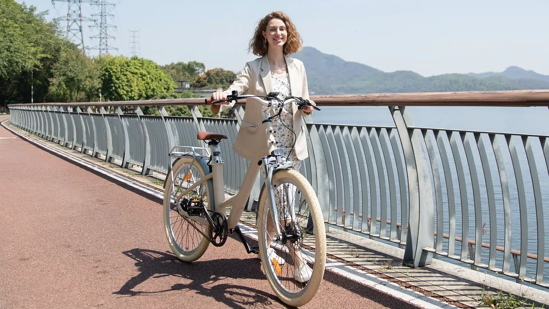 Eine Frau posiert lächelnd neben einem cremefarbenen ADO Air 28 Pro E-Bike. Sie steht auf einem roten Radweg, mit einem Geländer und einem See im Hintergrund.