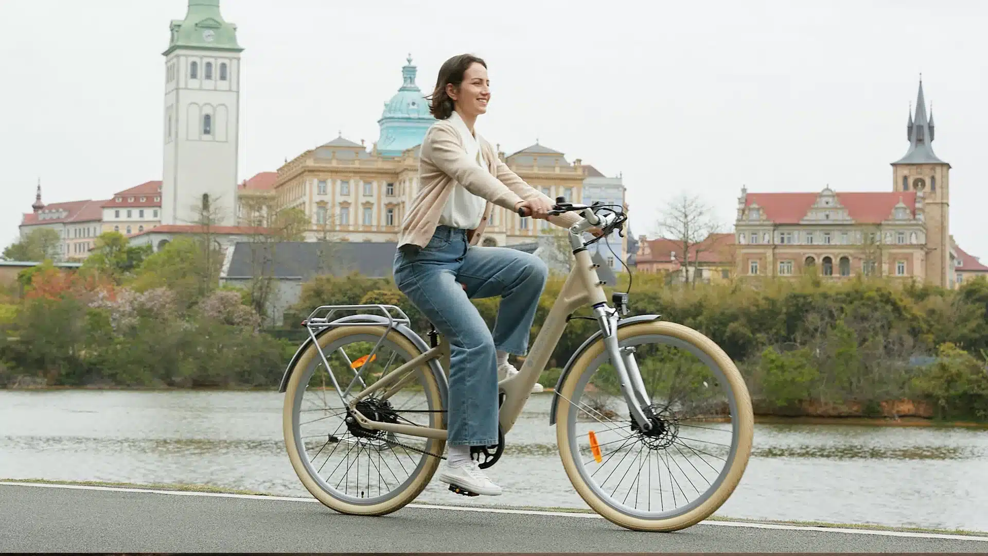 Eine Frau auf einem beigefarbenen ADO Air 28 Pro E-Bike mit Gepäckträger fährt auf einem asphaltierten Weg am Wasser. Im Hintergrund sind historische Gebäude mit Türmen und roten Dächern zu sehen.