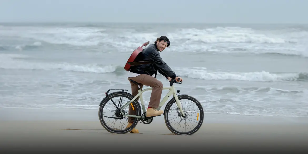 Mann mit Gitarre auf dem Rücken fährt ein beiges ADO Air 30 Pro | Ultra E-Bike am Strand, die Wellen sind im Hintergrund zu sehen.