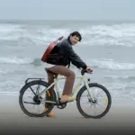 Mann mit Gitarre auf dem Rücken fährt ein beiges ADO Air 30 Pro | Ultra E-Bike am Strand, die Wellen sind im Hintergrund zu sehen.