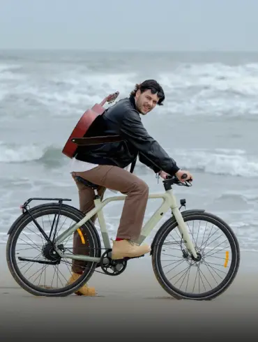 Mann mit Gitarre auf dem Rücken fährt ein beiges ADO Air 30 Pro | Ultra E-Bike am Strand, die Wellen sind im Hintergrund zu sehen.