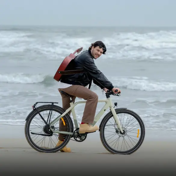 Mann mit Gitarre auf dem Rücken fährt ein beiges ADO Air 30 Pro | Ultra E-Bike am Strand, die Wellen sind im Hintergrund zu sehen.