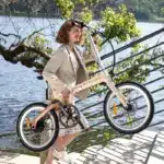 Frau mit beigem Blazer und Kleid trägt ein ADO Air Carbon Faltrad im Freien neben einer Treppe und einem Fluss.