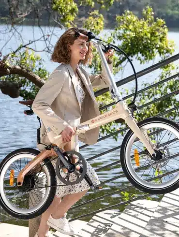 Frau mit beigem Blazer und Kleid trägt ein ADO Air Carbon Faltrad im Freien neben einer Treppe und einem Fluss.