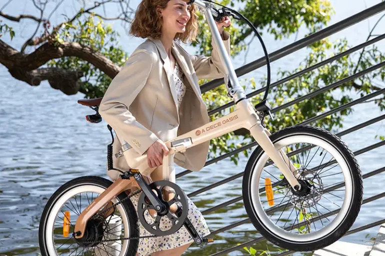 Frau mit beigem Blazer und Kleid trägt ein ADO Air Carbon Faltrad im Freien neben einer Treppe und einem Fluss.