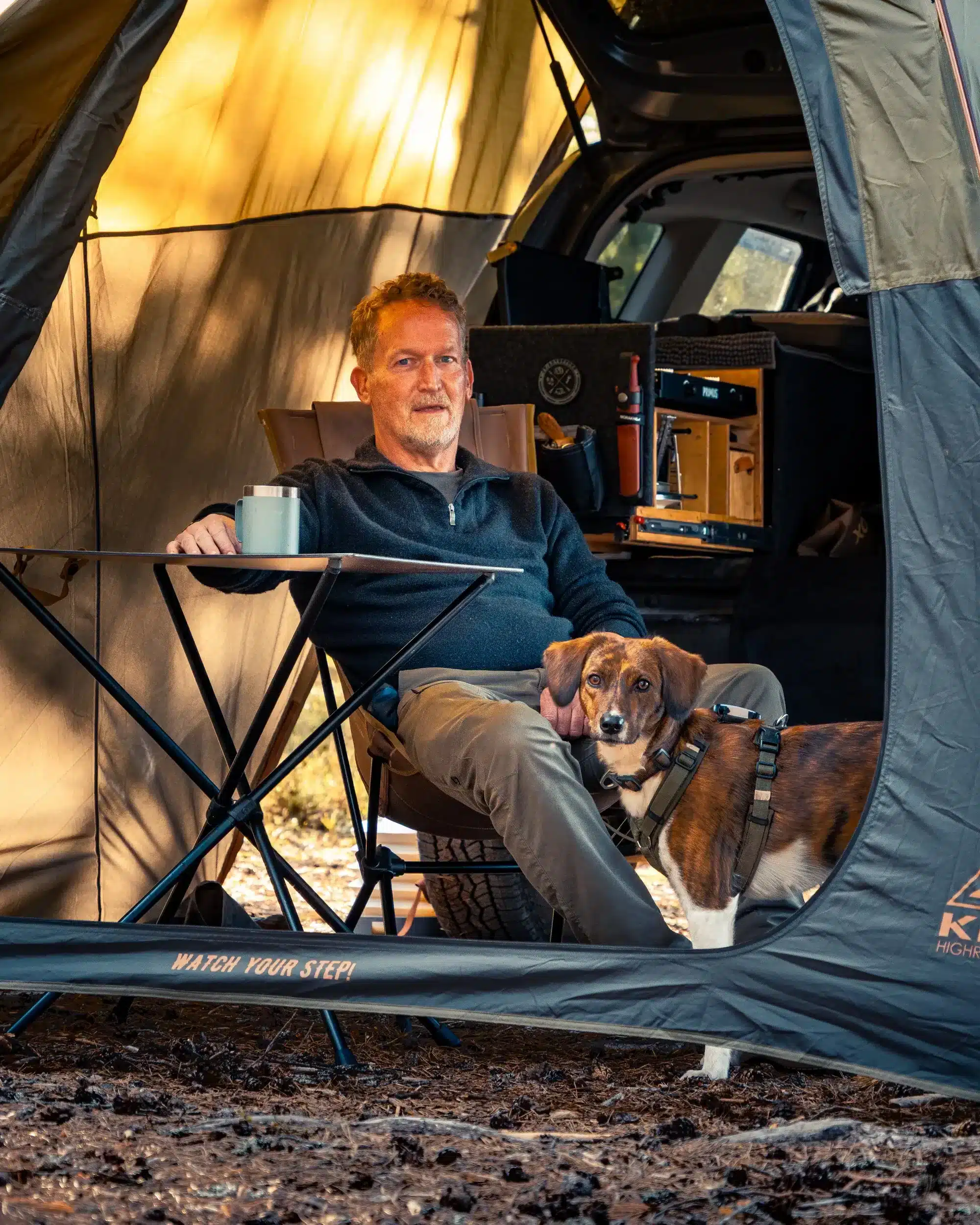Mann sitzt mit seinem Hund im Dachzelt neben einem Camper