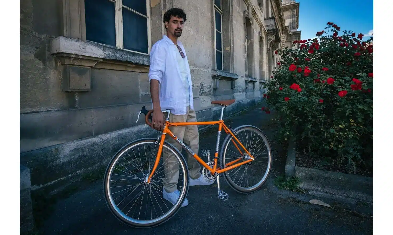 Mann mit orangefarbenem Stahlrahmen-Fahrrad mit braunem Lenkerband und braunem Sattel, stehend vor einer grauen Steinfassade und einem Busch mit roten Rosen.