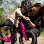 Eine Mutter beugt sich zu ihrem Kind, das auf einem pinken Motobécane Roadie Laufrad sitzt und hilft ihm.