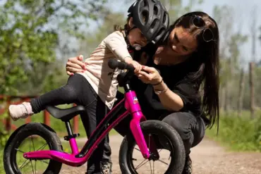 Eine Mutter beugt sich zu ihrem Kind, das auf einem pinken Motobécane Roadie Laufrad sitzt und hilft ihm.