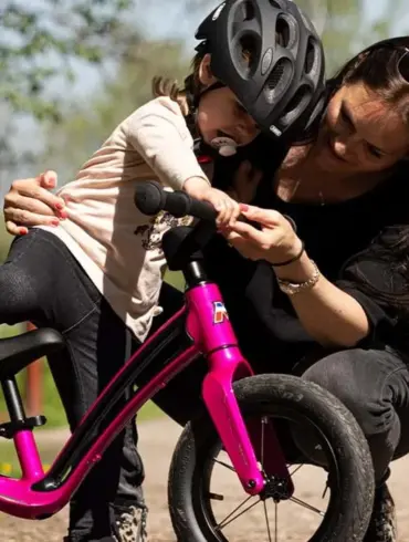 Eine Mutter beugt sich zu ihrem Kind, das auf einem pinken Motobécane Roadie Laufrad sitzt und hilft ihm.