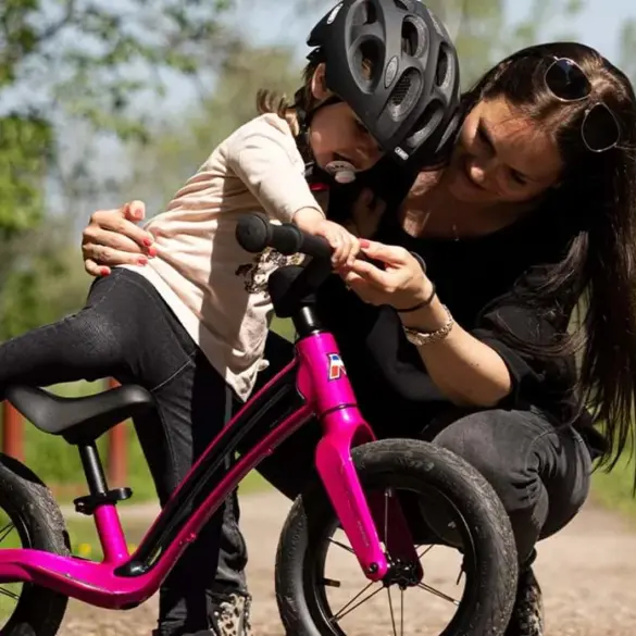 Eine Mutter beugt sich zu ihrem Kind, das auf einem pinken Motobécane Roadie Laufrad sitzt und hilft ihm.