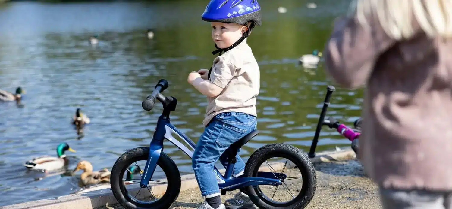 Kind mit Helm fährt ein blaues Laufrad mit 12-Zoll-Rädern am Ufer eines Sees.