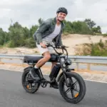 Mann in Jeansjacke und Shorts fährt ein schwarzes ENGWE M1 E-Bike mit breiten Reifen und Doppelsitz auf einer Straße.