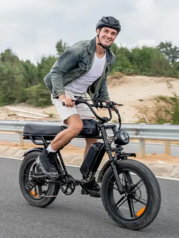 Mann in Jeansjacke und Shorts fährt ein schwarzes ENGWE M1 E-Bike mit breiten Reifen und Doppelsitz auf einer Straße.